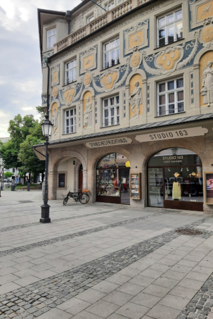 Auf diesem Foto sieht man den Blick auf eine Seite der Fassade des Ruffinihaus. Die Fassade ist mit Stuck verziert und zeigt Personen, Wappen und Muster. Außerdem sind Fenster in der Fassade. Die Farben sind gräulich/grün, blau und gelb/orange. Im Erdgeschoss sind zwei Geschäfte nebeneinander zu sehen. Links im Erdgeschoss ist ein Bogendurchgang zu sehen. Links im Bild neben der Fassade ist weiter weg ein Baum zu erkennen. Vor dem Haus stehen eine Laterne und ein Fahrrad, links im Bild.