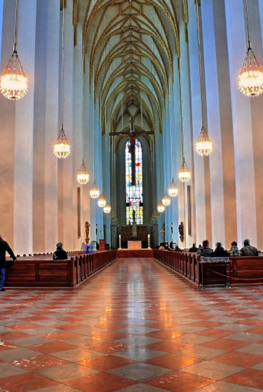 Auf diesem Foto ist die Innenansicht einer Kirche zu sehen. Die Decken sind sehr hoch. Links und rechts sind weiße Säulen zu sehen, die bis zur Decke ragen. Davor hängen links und rechts in einer Reihe nach vorne Lampen von der Decke. Darunter sind links und rechts Bänke in einer Reihe nach vorne von hinten fotografiert. Dazwischen führt ein breiter Gang nach vorne zum Altar. Hinter dem Altar ist ein großes Fenster zu erkennen.