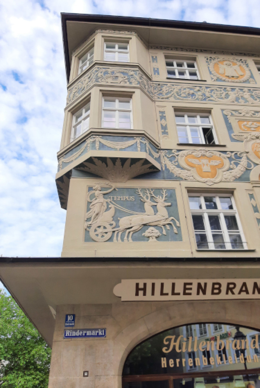 Auf diesem Foto sieht man eine nähere Aufnahme der Fassade. Das Foto wurde von unten hoch fotografiert. Man blickt also von der Straße auf die Fassade von unten. Man sieht eine Ecke der Fassade mit einem Erker. Die Fassade ist mit Stuck verziert, und man kann verschiedene Szenen, Wappen und Muster erkennen. Außerdem sind Fenster in der Fassade zu sehen. Darunter ist auf der Fassade „10, Rindermarkt“ als Straßennamen und Hausnummer zu lesen. Rechts im Bild unter der stuckverzierten Fassade erkennt man noch den Teil eines Schaufensters und darüber den abgeschnittenen Schriftzug „HILLENBRAN“.