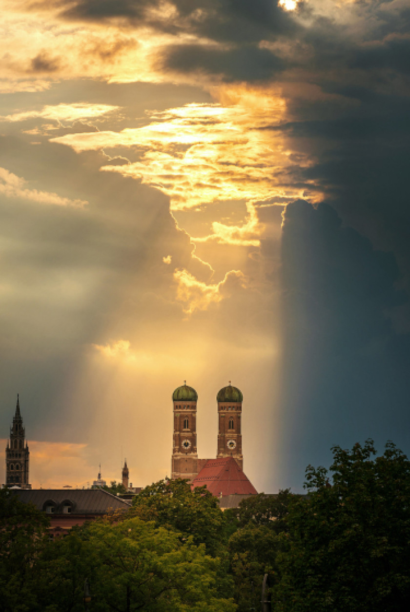 Auf diesem Foto sieht man in der Ferne zwei Türme, die von dem Sonnenlicht angestrahlt werden. Davor sind Bäume zu sehen. Am Himmel sind Wolken und an einer Stelle über den Türmen strahlt die Sonne durch die Wolken. Der Rest liegt im Schatten.