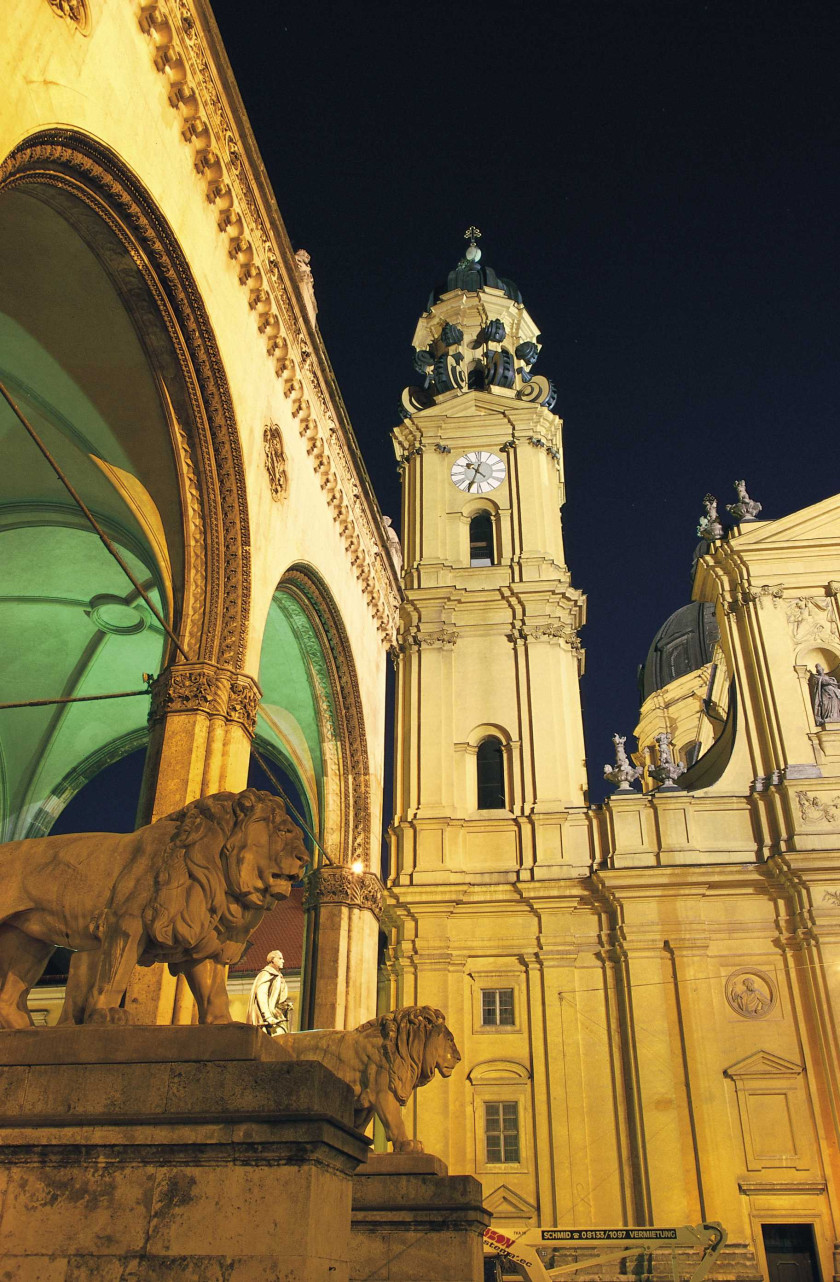 Auf diesem Foto sieht man einen Teil einer gelben Außenfassade einer Kirche. Links im Bild sieht man Statuen von zwei Löwen und eine Halle dahinter von der Seite aufgenommen. Die Halle ist innen grün beleuchtet und man sieht durch große Bögen einen Teil der Decke. Im Zentrum und im rechten Teil des Bildes sieht man einen gelben Turm an der Außenfassade einer Kirche, die nur zu einem kleinen Teil erkennbar ist. Der Himmel darüber ist schwarz. Das Foto ist eine Nachtaufnahme.