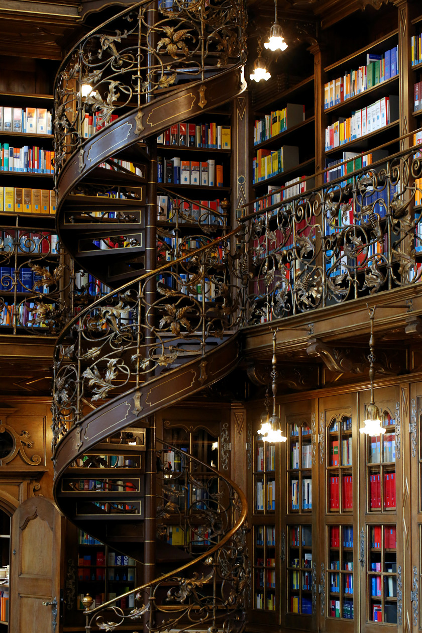 Auf diesem Foto sieht man eine Wendeltreppe im Fokus. Dahinter sind gefüllte Bücherregale auf zwei Ebenen zu sehen.