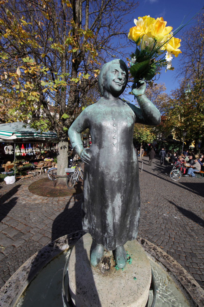 Auf diesem Foto sieht man die Skulptur einer Frau im Zentrum. Die Frau steht auf einem Sockel aus Stein in einem Brunnen und soll Liesl Karlstadt darstellen. In der einen Hand hält sie einen gelben Blumenstrauß hoch neben ihr Gesicht. Im Hintergrund ist der Viktualienmarkt zu erkennen.