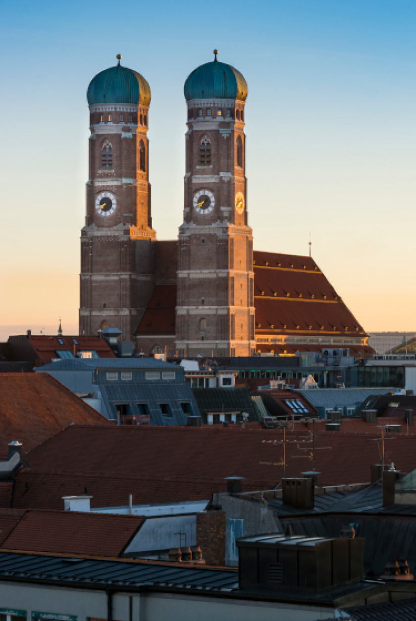 Auf diesem Foto sieht man zwei Türme an einer Kirche im Zentrum des Fotos. Im Vordergrund sind im Schatten Dächer von Häusern zu sehen. Mittendrin bzw. hinter den Dächern steht die Kirche mit zwei großen Türmen.