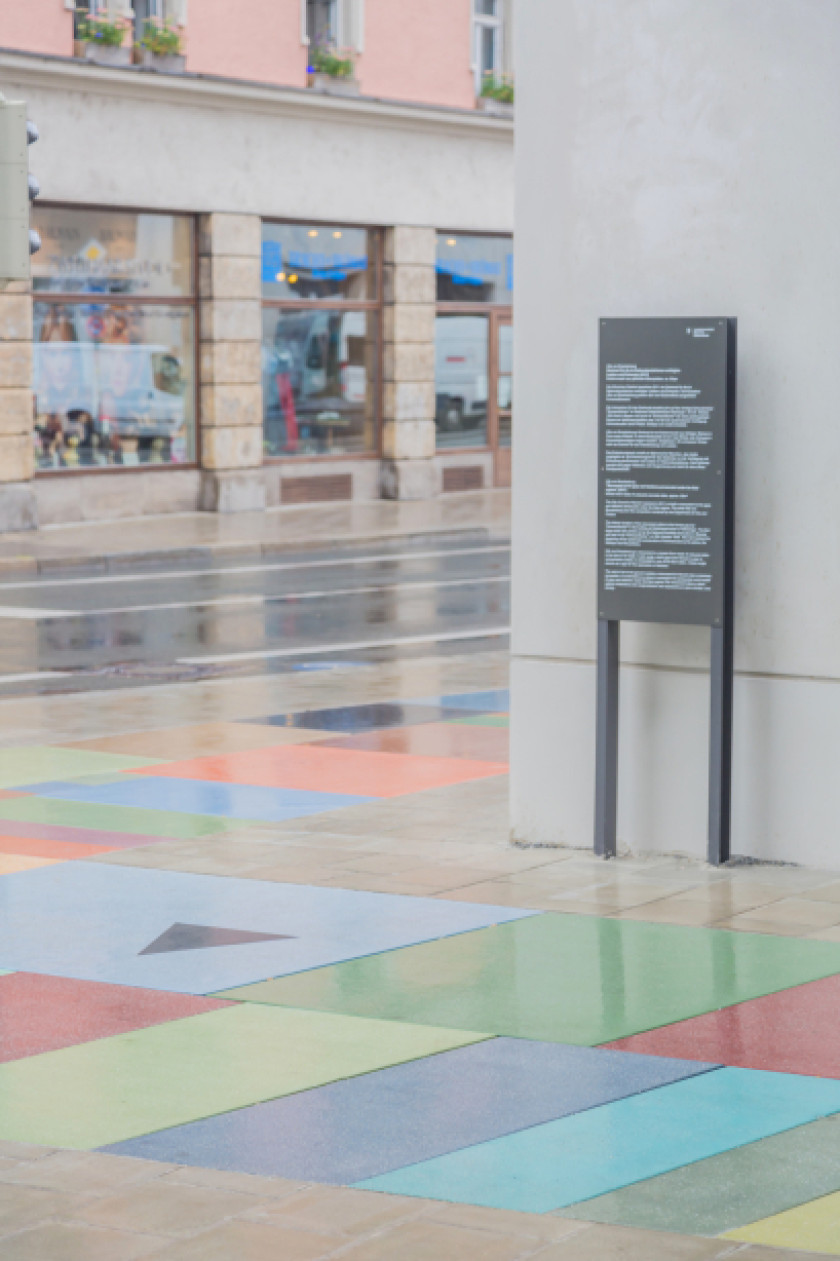 Auf diesem Foto sieht man bunte Bodenfliesen von seitlich oben fotografiert. Dahinter ist an der Wand eine Informationsstele. Links im Hintergrund sieht man Schaufenster von Geschäften.