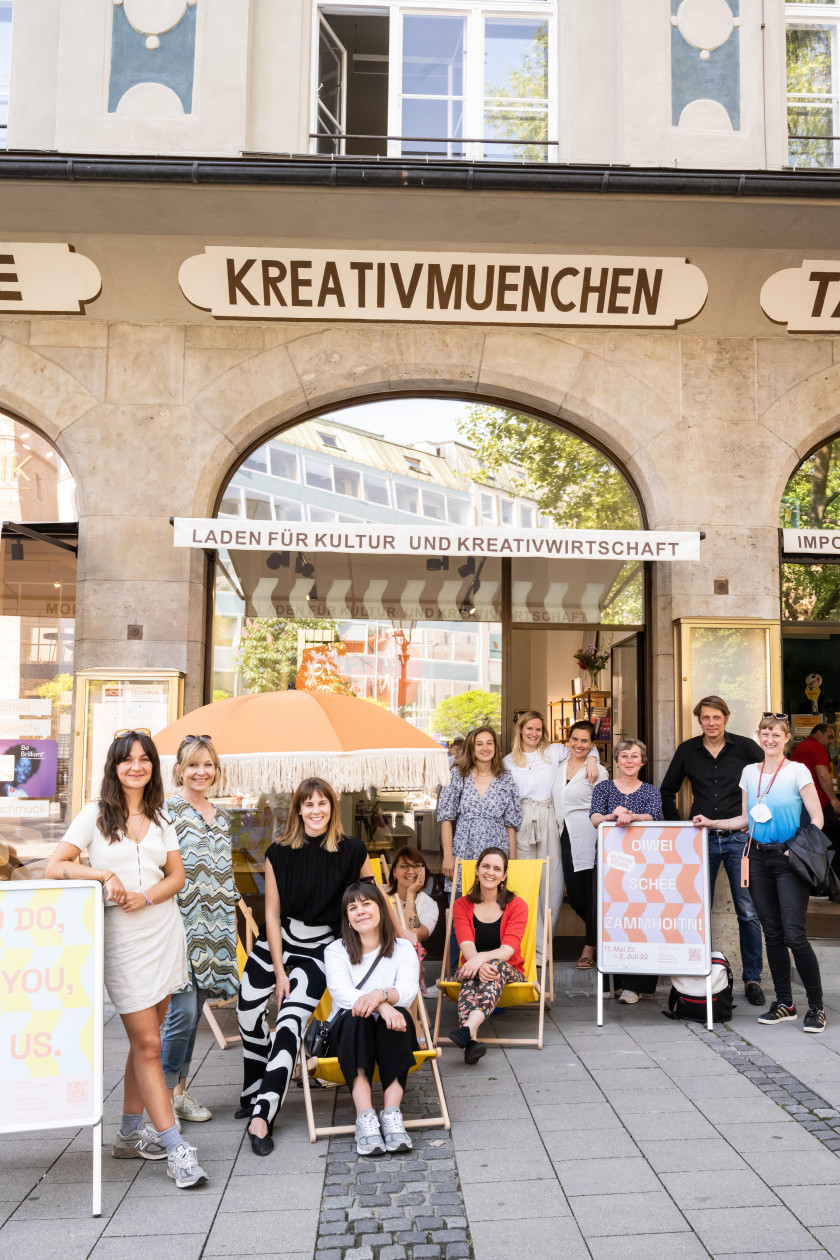 Auf diesem Foto sieht man verschiedene Personen, die vor einem Geschäft stehen und in die Kamera blicken. Manche stehen und manche sitzen auf Liegestühlen. Sie sind vor einem Geschäft mit dem Schriftzug „KREATIVMUENCHEN“ und „Laden für Kultur und Kreativwirtschaft“.