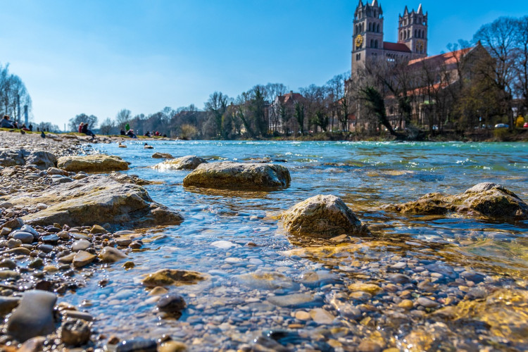 The picture shows the church of St. Maximilian with its striking towers jutting out from behind bare trees. In the foreground, the stony riverbank glistens in the clear water, while the stony riverbed shimmers through in different colors.
